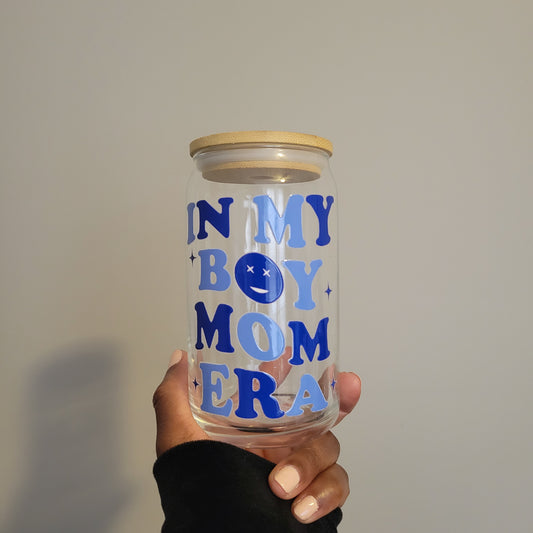  Holding a 16oz glass cup from SuppliedByKai featuring a UV DTF design with the phrase 'In My Boy Mom Era' in dark and light blue lettering, accompanied by a cheerful smiley face, complemented by a bamboo lid