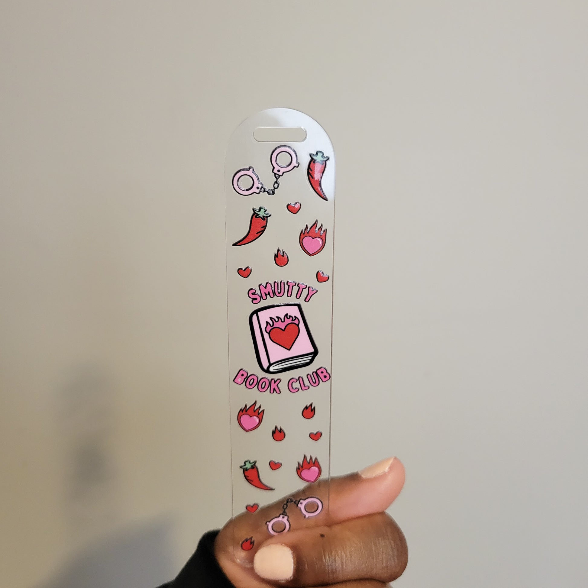 Hand holding the Smutty Book Club acrylic bookmark by Supplied By Kai, featuring UV DTF print with pink and red spicy-themed designs, including chili peppers, pink handcuffs, flames, and hearts on a clear acrylic background.