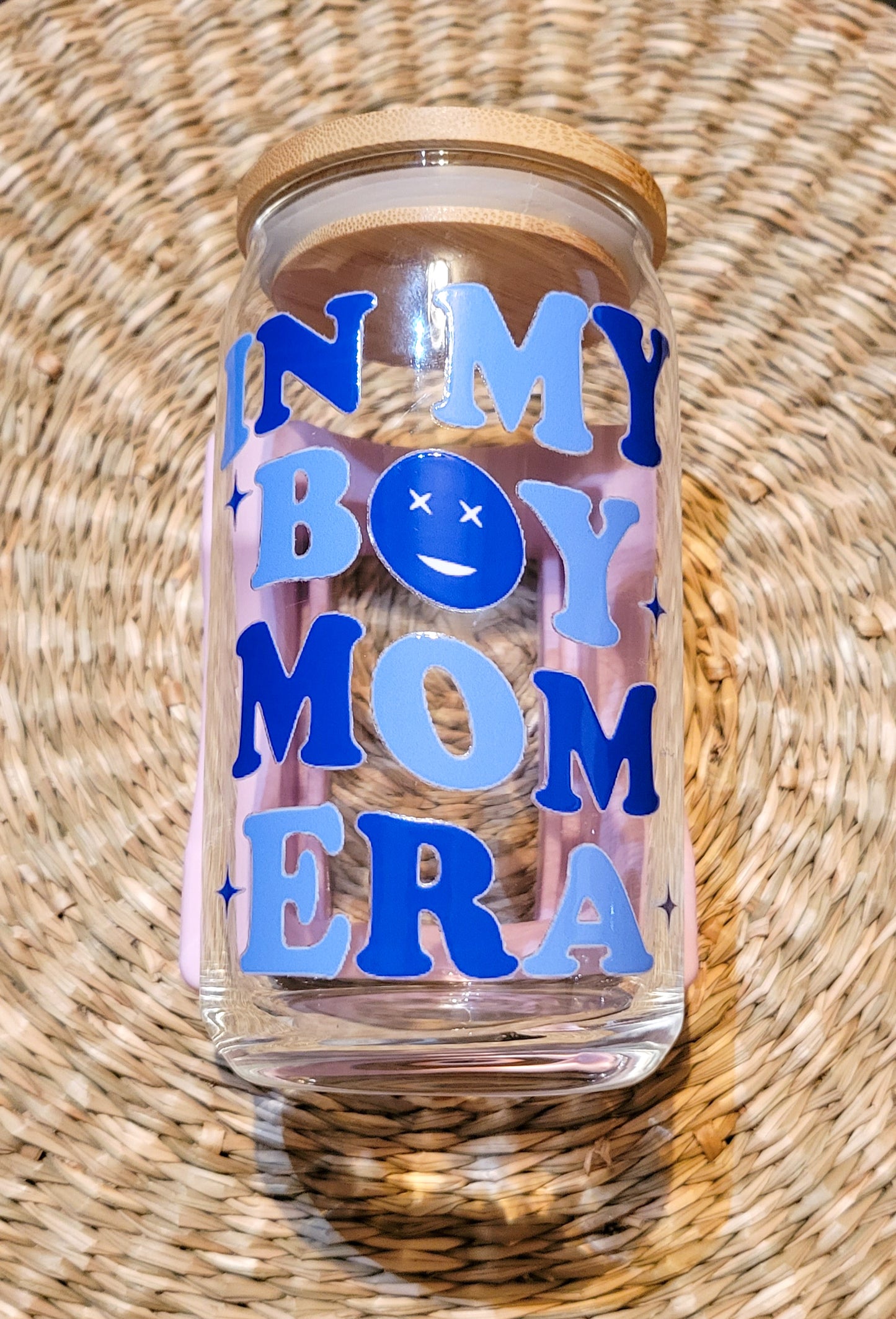 A close-up of a 16oz glass cup from SuppliedByKai featuring a UV DTF design with the phrase 'In My Boy Mom Era' in dark and light blue lettering, accompanied by a cheerful smiley face, complemented by a bamboo lid placed on a round woven seagrass placemat