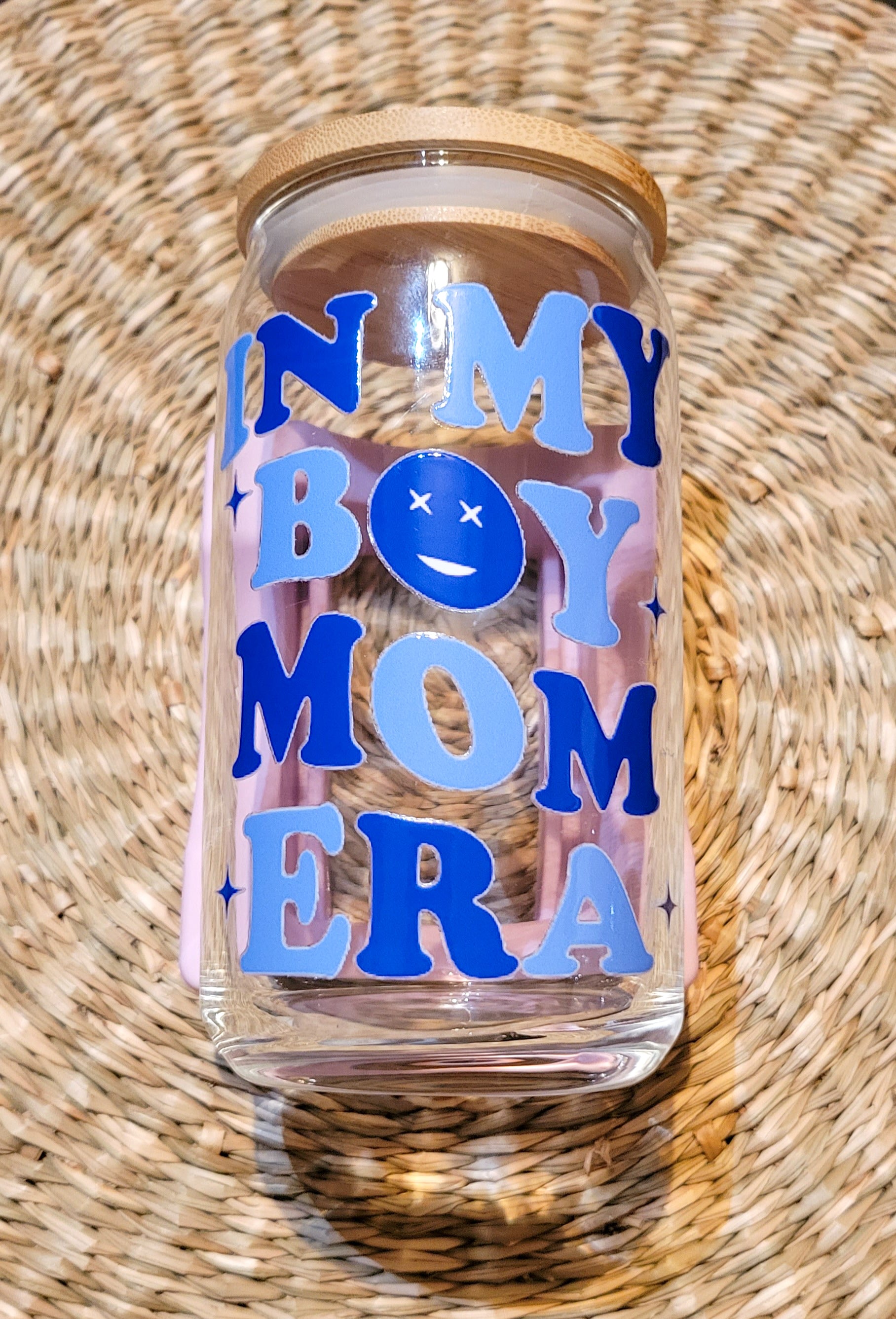 A close-up of a 16oz glass cup from SuppliedByKai featuring a UV DTF design with the phrase 'In My Boy Mom Era' in dark and light blue lettering, accompanied by a cheerful smiley face, complemented by a bamboo lid placed on a round woven seagrass placemat