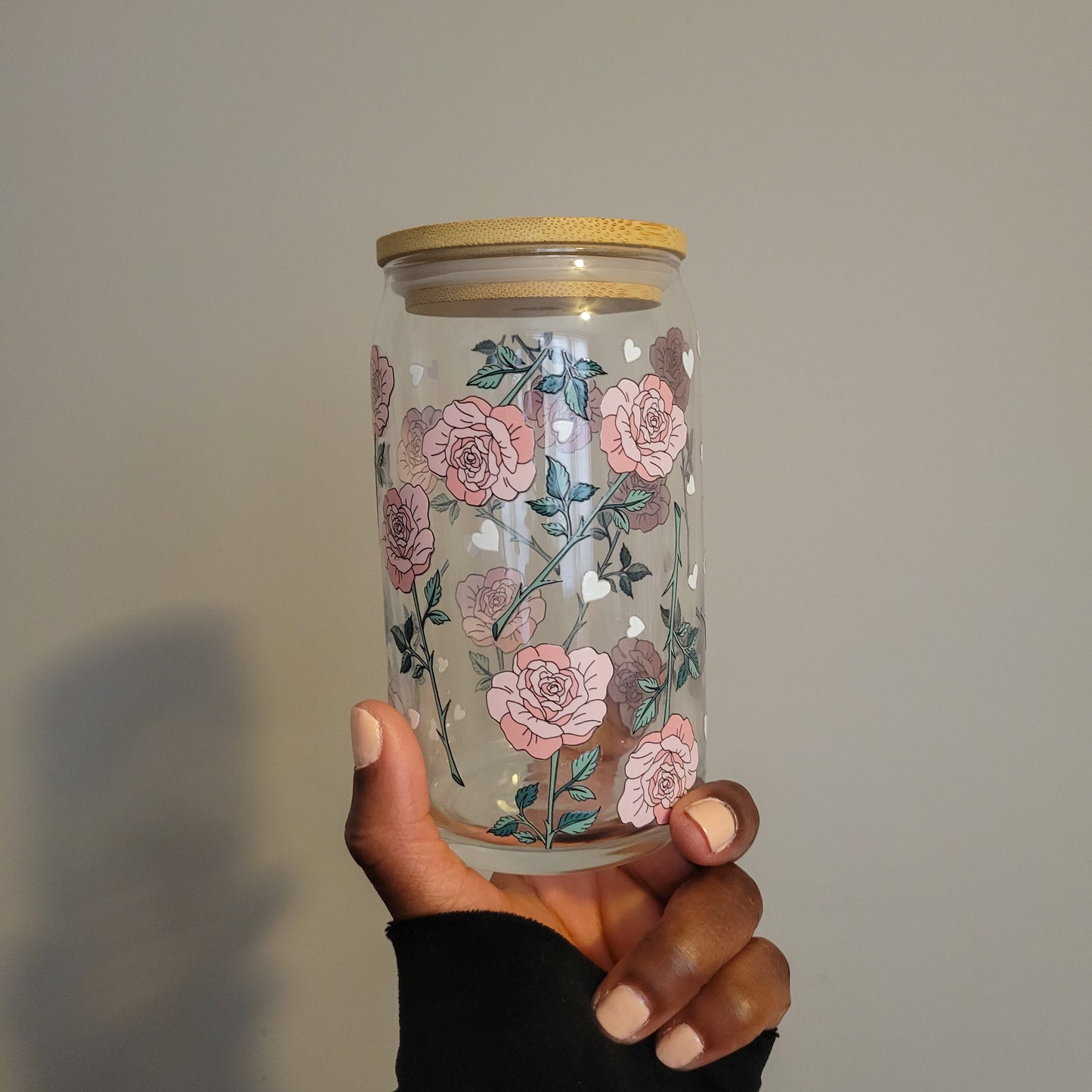 A person holding a 16oz Glass Cup styled like a glass beer can, featuring a double-sided UV DTF design of soft pink roses with dark green thorns and small white hearts. 16oz glass cup has a bamboo lid perfect for an elegant and stylish drinkware option from SuppliedByKai