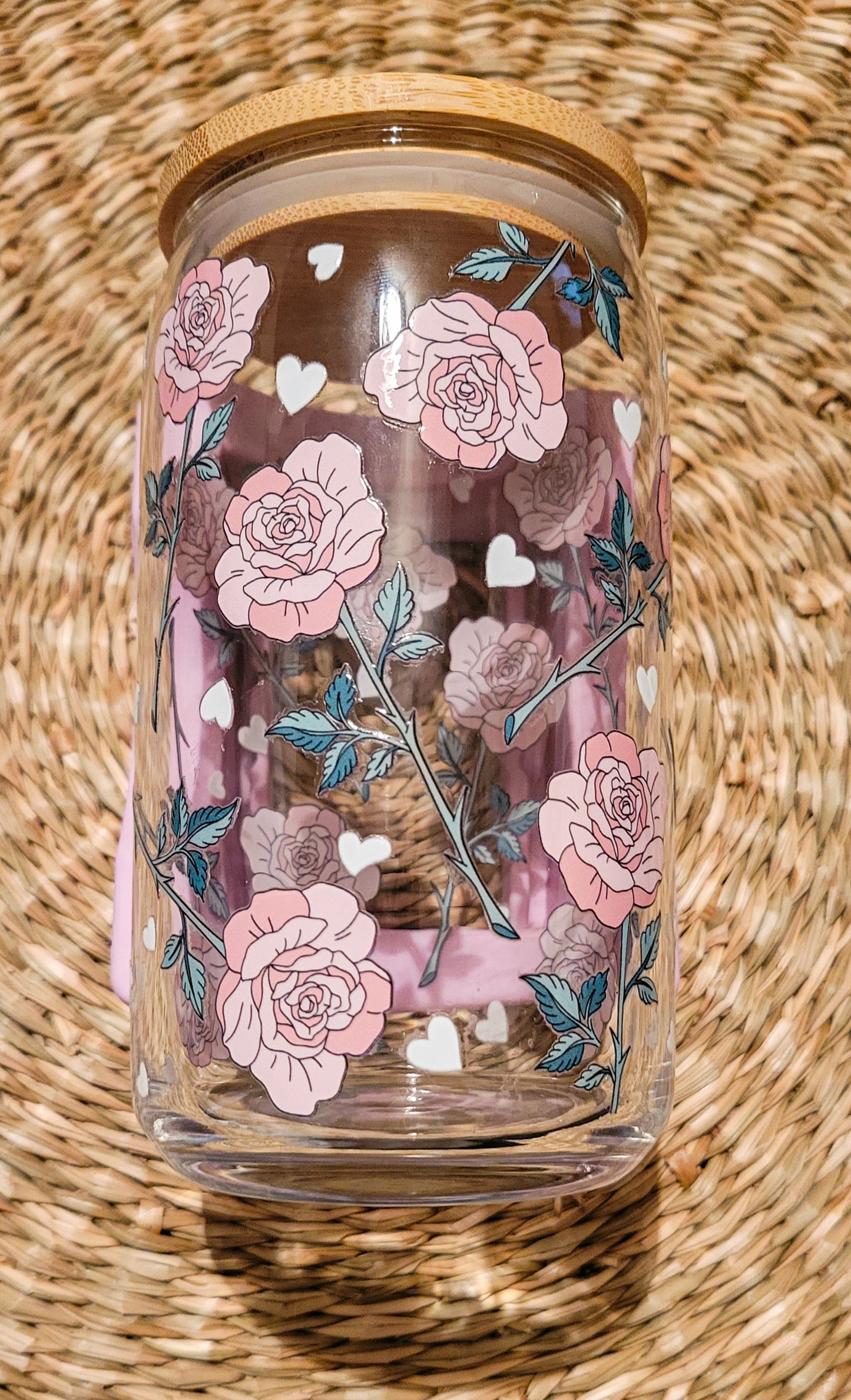 A 16oz glass can cup with a bamboo lid showcasing a UV DTF pink roses floral design with small white heart, elegantly placed round woven seagrass placemat. 