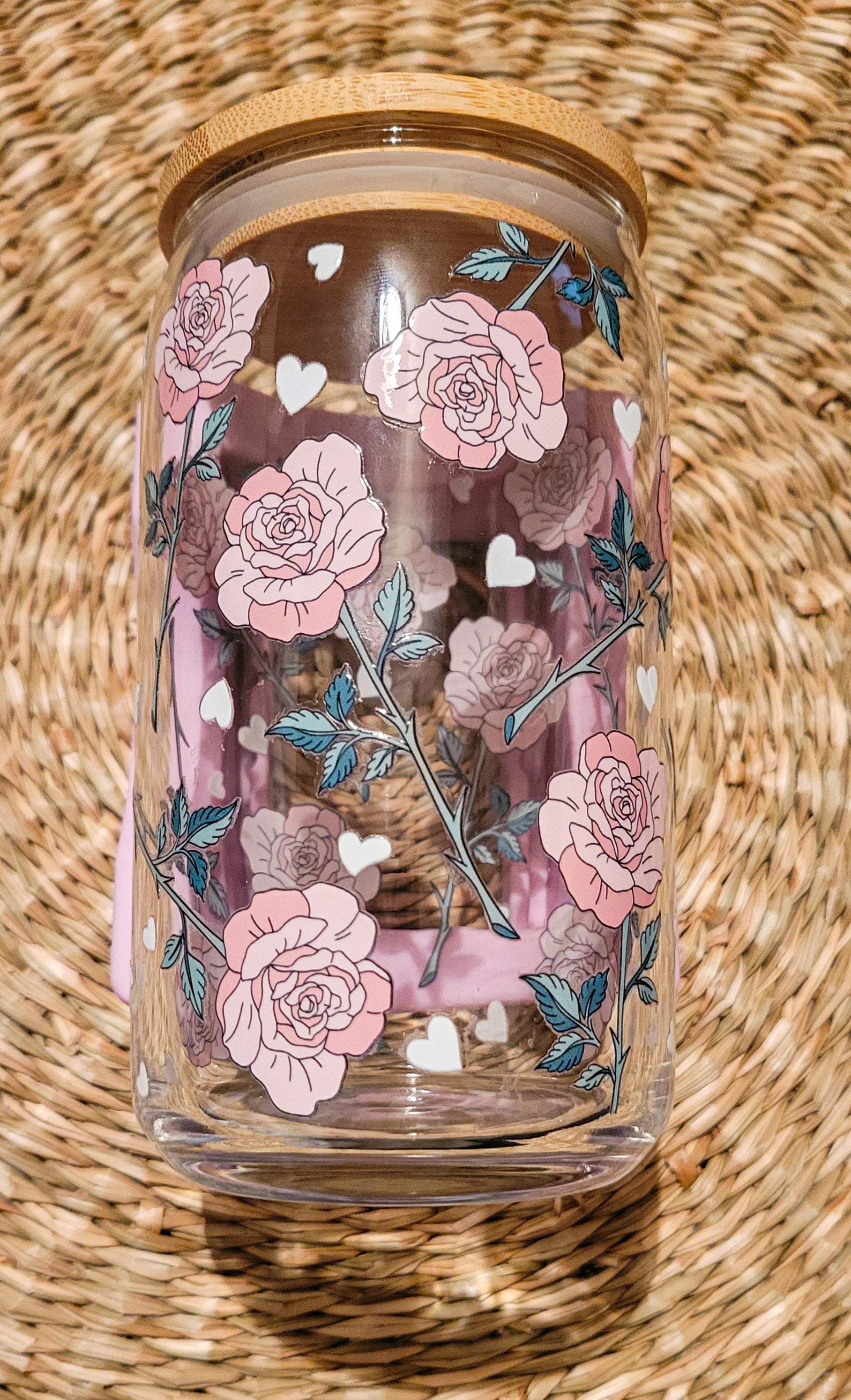 A 16oz glass can cup with a bamboo lid showcasing a UV DTF pink roses floral design with small white heart, elegantly placed round woven seagrass placemat. 